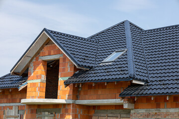 Unfinished residential house construction showing red brick walls and new dark metal roof tiles. A modern skylight shines from the building structure.