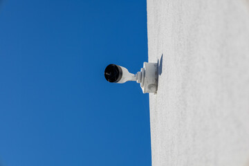 Fototapeta premium Modern security camera monitoring an outdoor area. White surveillance device fixed to a textured wall under a bright blue sky.