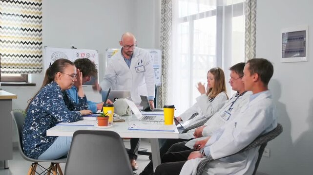 Senior doctor in white coat angrily criticizes interns during a clinical meeting. Group of medical workers looks stressed and thoughtful while listening to strict remarks about mistakes in research.
