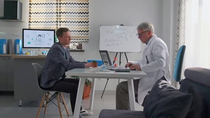 Senior doctor in lab coat consulting a man in business suit in modern medical office. They discuss medical notes, scientific research and possible treatment options. - Powered by Adobe