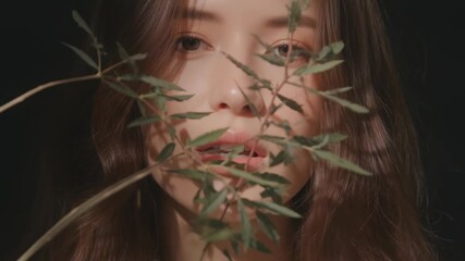 Close-up portrait of a young woman partially hidden behind delicate green leaves, natural lighting