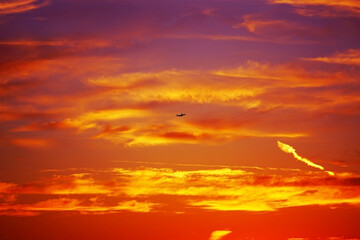 Naklejka premium Dramatic sky at sunset with airplane silhouette flying through colorful clouds. Fiery tones of orange, yellow and red blend creating breathtaking view. Concept of travel, freedom, wanderlust, aviation