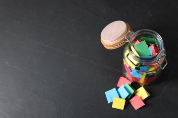 Colorful paper pieces in glass jar on black table, above view. Space for text. Notes with messages