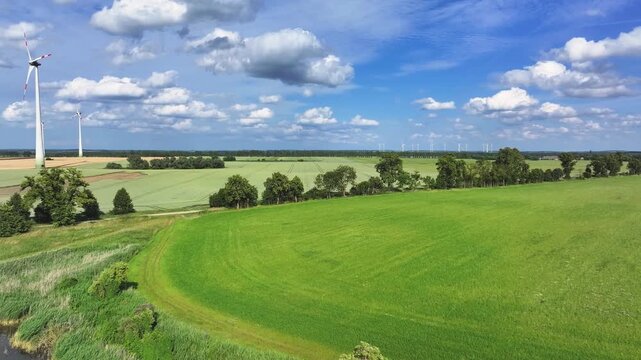 Aerial view of towering wind turbines standing amidst vibrant green fields under a blue sky dotted with fluffy white clouds, Br&uuml;ssow Uckermark, Brandenburg, Germany.
