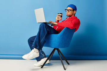 Trendy young man in beanie works on laptop while sipping coffee against blue backdrop