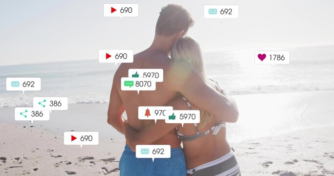 Hugging couple wearing bikini top and swim shorts at sandy beach, with floating social media icons