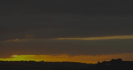 Glowing yellow orange horizon strip spanning rolling hills in countryside at dusk, moody cloudy sky