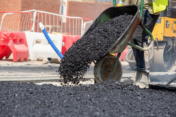 Team of road workers in bright orange uniforms repair road surface with wheelbarrow filled with hot asphalt