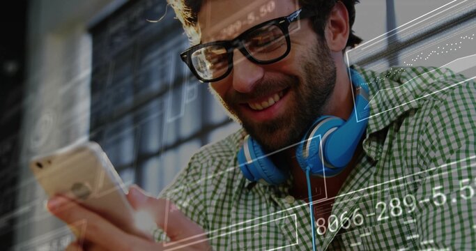 Smiling man wearing glasses and blue headphones analyzing smartphone in workspace, data overlays