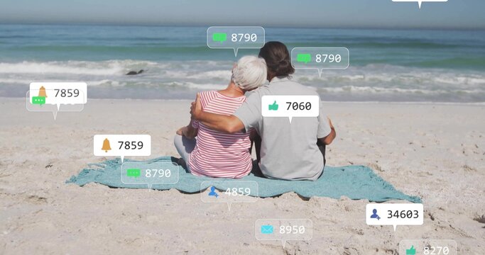 Embracing senior couple sitting on blue towel watching waves at shoreline, with social media icons
