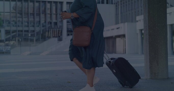 Walking woman in teal dress pulling suitcase at building entrance, with bag, coffee cup, copy space