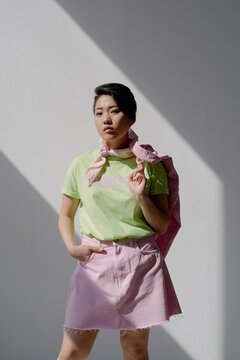 Portrait of young adult Asian woman standing with hand in pocket, wearing casual outfit and scarf around neck, looking directly at camera with confident expression, sunlight casting shadows