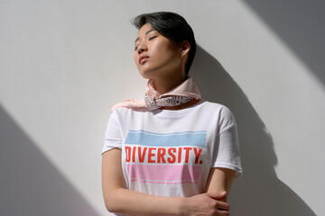 Young adult Asian woman standing with eyes closed leaning against wall wearing t shirt with diversity message arms crossed across chest sunlight casting shadows