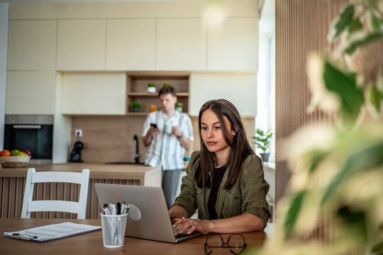 Focused woman working remotely while man using smartphone in background