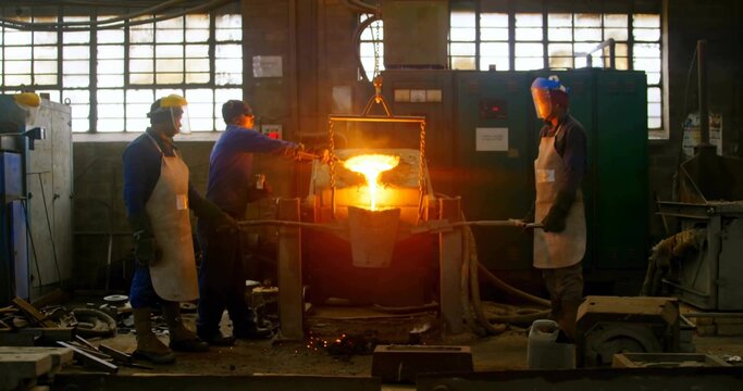 Pouring glowing metal from suspended crucible into mold in foundry wearing protective coveralls