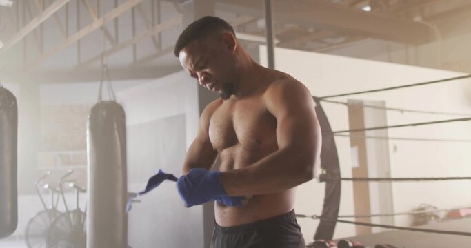 Wrapping hands, muscular boxer tightening blue hand wraps in boxing ring, with punching bag