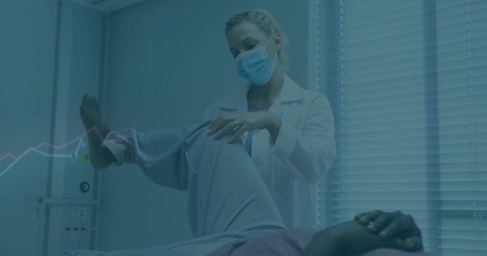 Lifting patient leg on exam table, doctor wearing coat mask assessing knee motion in exam room