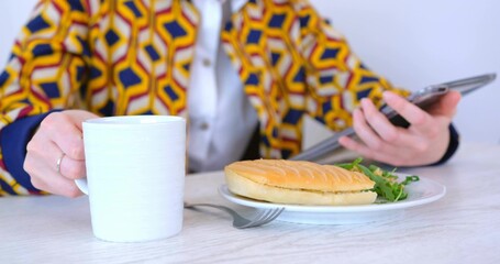 Holding white mug scrolling tablet wearing printed jacket at cafe table with panini fork and greens