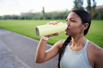 Young female athlete enjoying a refreshing drink from a reusable bottle after an intense workout in...