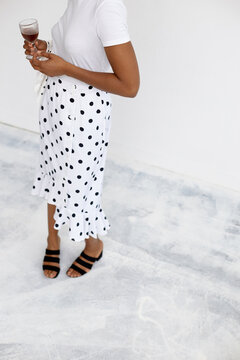 Unrecognizable Black woman standing holding wine glass, wearing polka dot skirt and sandals, upper body partially out of frame, posing against light background