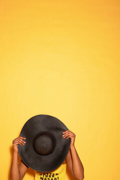 Black woman holding large black hat in front of face against yellow background, only upper body and hands visible, creating playful and mysterious pose with ample copy space