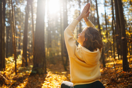 Relaxed woman practicing yoga poses, spreading arms in sunny autumn forest. Young woman enjoying weather surrounded by colorful leaves and doing breathing exercises. Relaxation, rest concept.