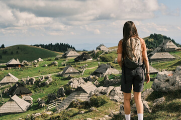 Naklejka premium Hiker pauses, gazing at a picturesque alpine village nestled amongst rolling green hills and rocky terrain. A stunning view of traditional wooden houses under a cloudy sky.