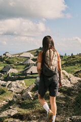 Hiker pauses, gazing at a picturesque alpine village nestled amongst rolling green hills and rocky...