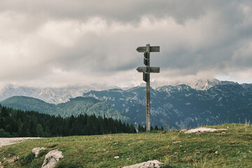 Wooden signpost on a grassy hilltop overlooking majestic mountains under a cloudy sky.  A scene of natural beauty and contemplation, perfect for travel, adventure, or nature-themed projects.