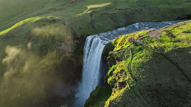 Golden hour aerial footage of Skogafoss waterfall, Iceland. The tall cascade flows between lush cliffs while glowing in warm evening light, filmed in cinematic 4K drone ProRes.