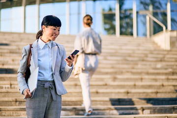 Fototapeta premium Asian Businesswoman Viewing Smartphone While Carrying Laptop on City Stairs