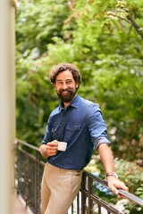Smiling Man Holding Coffee Cup on Outdoor Balcony with Greenery Background
