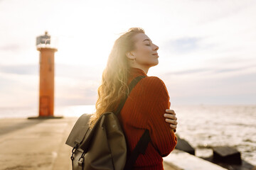 Happy woman having fun on the pier with a lighthouse. Tourist with a backpack enjoying the seascape...