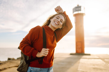 Happy woman having fun on the pier with a lighthouse. Tourist with a backpack enjoying the seascape...