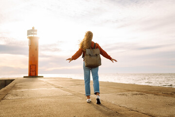 Happy woman having fun on the pier with a lighthouse. Tourist with a backpack enjoying the seascape...