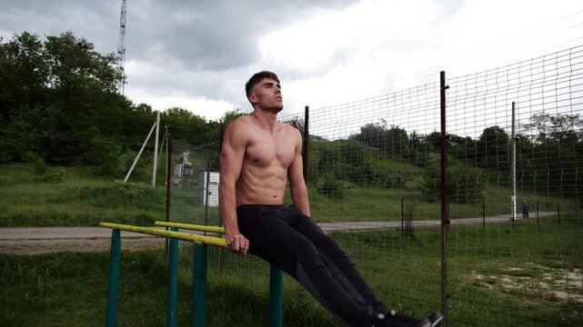Strong man trains abdominal muscles raising legs on parallel bars at sports ground. Young shirtless sportsman exercising at nature. Sporty athlete working out at nature. Concept of sportive lifestyle