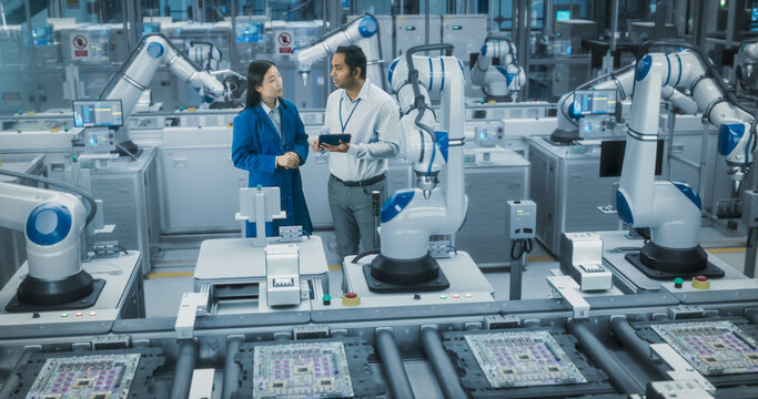 Electronics Factory Floor: Female Project Manager and Engineer Talking, Using Tablet Computer. Monitoring Advanced Equipment Production. Robotic Arms Assembly Line Manufacturing Circuit Boards