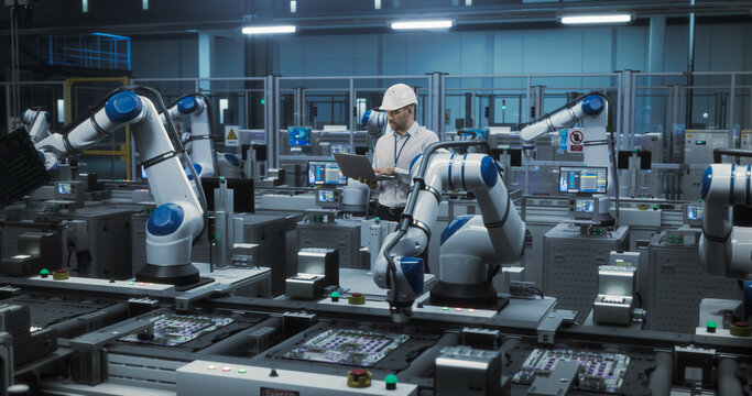 Portrait of a Caucasian Engineer in a Hard Hat Working in an Industrial Facility. Industrial Manager Using Laptop Computer to Monitor Robotic Arms in a Futuristic Electronics Factory