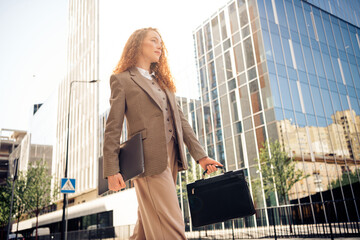 Confident female businesswoman walking outdoors holding a briefcase while wearing a formal suit and...