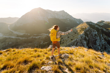 Female hiker with hiking backpack stands on top of mountain, admiring mountain landscape at sunset. Traveler feels freedom, enjoys the sun's rays in the mountains. Adventure concept, nature.