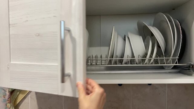person's hand opens a modern, light-colored wooden kitchen cabinet, showing clean white plates and bowls neatly arranged on a metal dish drying rack. A concept of kitchen storage and home organization