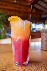 Refreshing tropical cocktail, layered red and orange drink in tall glass on wooden table