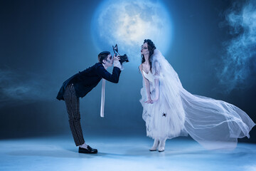 Gothic couple surreal photoshoot with vintage camera and playful pose under blue moon. Concept of Halloween fashion, bizarre editorial art, creative album cover, and dramatic theatrical aesthetic.