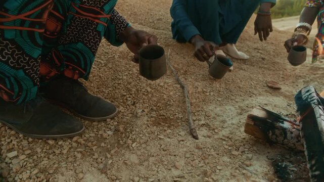 Group of three Black friends in ethnic clothes sitting in front of campfire, pouring some coffee on sandy ground then drinking beverage