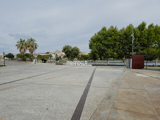The open square by the Tua River in Mirandela, Portugal, is framed by palm trees and city landmarks. Inviting for gatherings, this riverside space connects people to nature and urban charm.