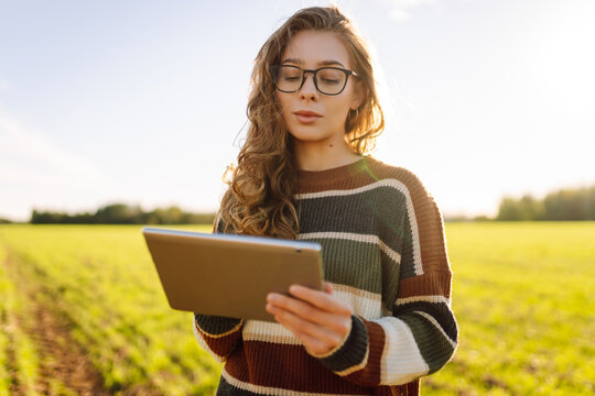 Young female farmer with digital tablet in green agricultural field in sun. Female agronomist in field with green sprouts checks the growth and quality of the crop. Farming and technology concept. - Powered by Adobe
