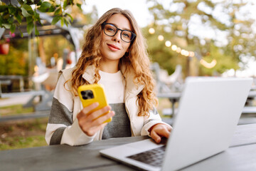 Young woman in glasses with a laptop sitting at a wooden table in the park. Beautiful female...