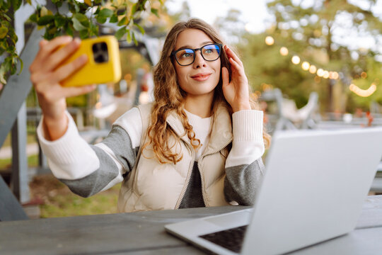 Young woman in glasses with a laptop sitting at a wooden table in the park. Beautiful female freelancer with a phone enjoying nature in the autumn forest. Remote work and nature concept.