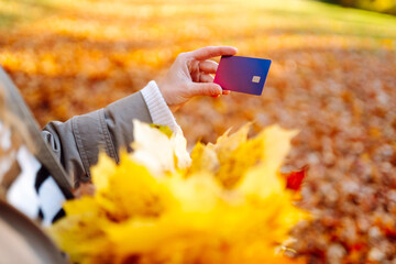 Close-up of young woman with bouquet of autumn leaves holding bank card in hand at sunset. A woman with debit card against the backdrop of an autumn sunny park. Concept of technology, walking.