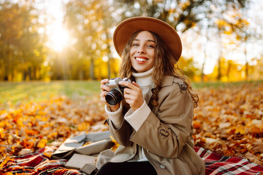 Woman is sitting on blanket in park covered with autumn leaves with camera in hands. Beautiful woman in hat is enjoying autumn landscape and taking pictures on camera. Concept of beauty, relaxation.